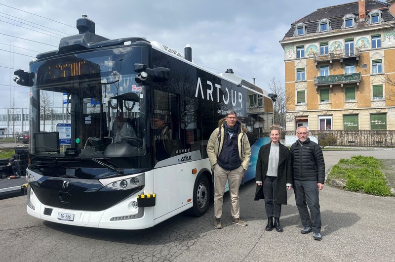 Drei Personen stehen vor einem weißen Bus mit der Aufschrift ARTOUR auf der Seite vor einem mehrstöckigen Wohnhaus.